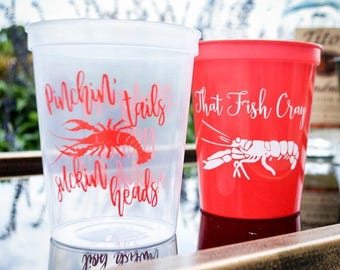 May include: A translucent plastic cup with red text reading Pinchin' tails and suckin' heads above a red crawfish graphic, next to a solid red plastic cup with white text reading That Fish Cray above a white crawfish graphic.