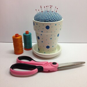 May include: A white flower pot pin cushion with blue polka dots and a blue and white houndstooth cushion. Red-tipped pins are inserted into the cushion. Pink and black scissors and two spools of thread are also present.