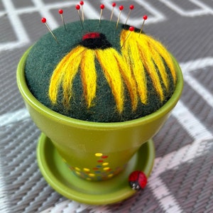 May include: A handmade pincushion in a green ceramic pot, featuring a felted flower design with yellow petals and a black center. Red-headed pins are inserted into the green felt. A small ladybug decorates the pot's edge.