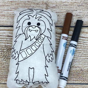 Op de afbeelding: Een wit pluche speelgoed in de vorm van een stripfiguur, met zwarte gestikte contouren. Twee Crayola wasbare stiften, bruin en zwart, liggen naast het speelgoed op een houten oppervlak. Het speelgoed heeft een riem met rechthoekige details.