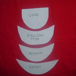 May include: A red fabric surface with four white paper templates. The templates are labelled "LONG", "BALLCAP style", "MEDIUM", and "SHORT". The curved shapes suggest a pattern for a hat or similar item.
