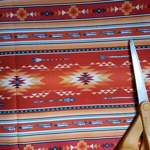 Puede incluir: Primer plano de una tela con un dise&ntilde;o de inspiraci&oacute;n nativa americana. La tela presenta un patr&oacute;n repetido de plumas, flechas y formas geom&eacute;tricas en tonos rojos, naranjas y turquesas. Un par de tijeras amarillas es parcialmente visible.