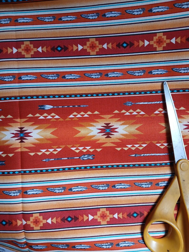 Puede incluir: Primer plano de una tela con un dise&ntilde;o de inspiraci&oacute;n nativa americana. La tela presenta un patr&oacute;n repetido de plumas, flechas y formas geom&eacute;tricas en tonos rojos, naranjas y turquesas. Un par de tijeras amarillas es parcialmente visible.