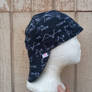 May include: Black welding cap with a white scientific formula print. The cap features equations, diagrams, and the text "PRISM" and "WHITE LIGHT". Includes a small American flag patch. The cap is designed to protect the head and neck.