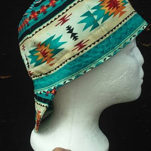 May include: A colorful, patterned welding cap with a southwestern design. The cap features a teal, cream, orange, and black geometric pattern. The cap is displayed on a white foam head against a dark background.