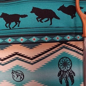 May include: Teal fabric with a Native American-inspired design. Features black silhouettes of wolves, dream catchers, and geometric patterns in shades of teal, tan, and red. An orange pair of scissors is partially visible.