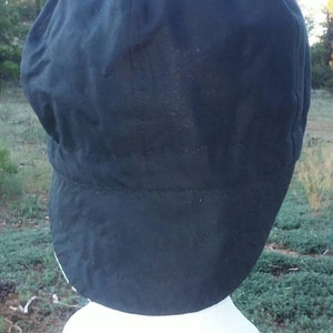 May include: A black newsboy cap with a rounded crown and a short, stiff brim. The cap is made of a dark fabric and is displayed on a white mannequin head. The background shows a natural outdoor setting.