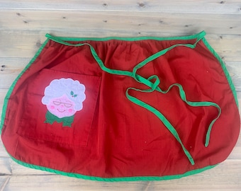 May include: A red waist apron featuring a green trim and long green waist ties. A square pocket on the left side displays an embroidered applique of a white-haired character with glasses, pink cheeks, and a green bow tie with holly leaves.