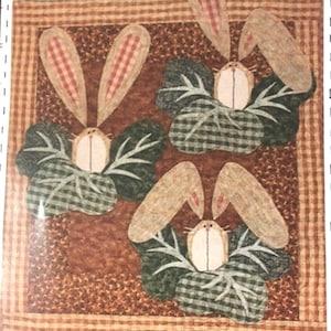 Puede incluir: Tres conejos marrones con orejas rosas asoman por detrás de plantas verdes con hojas. Los conejos están sobre un fondo a cuadros marrón y beige.