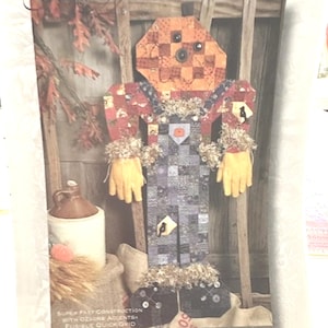 May include: A fabric scarecrow wall hanging featuring a patchwork pumpkin head with button eyes, a red patterned shirt, and grey checkered overalls. The figure has yellow hands, frayed cuffs, and decorative patches. Text at the bottom reads: SUPER FAST CONSTRUCTION WITH OZONE ACCENTS FUSIBLE QUICK GRID.