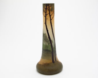 May include: A tall, slender vase with a landscape design. The vase features a scene of trees and a sunset in shades of brown, green, and yellow. The base is a darker brown, and the vase has a flared bottom. The artist's signature, "Legras", is visible.