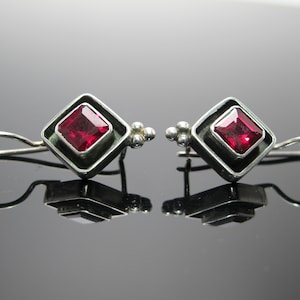 May include: A pair of silver earrings with a square, deep red gemstone set within a diamond-shaped frame. The earrings have a simple hook design and small, clustered silver accents.