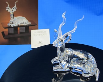 May include: A clear crystal gazelle figurine with detailed facets. The figurine has long, curved horns and is lying down. A certificate and a picture of the figurine are in the background. The figurine is on a black surface.