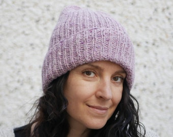 Könnte beinhalten: Eine Frau mit langen, dunklen, lockigen Haaren trägt eine weiche, lilafarbene Strickmütze. Sie schaut direkt in die Kamera und lächelt leicht.