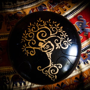 May include: A black handpan with a gold tree of life design. The handpan is sitting on a colorful fabric with an elephant pattern.
