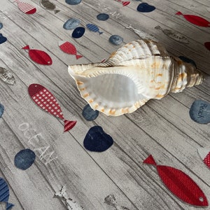 May include: A large, beige seashell with a spiral shape rests on a gray fabric. The fabric is patterned with red, blue, and white fish and pebbles, with the word "OCEAN" printed on it. The overall theme is nautical.