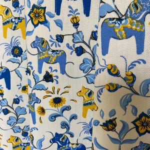Puede incluir: Tela blanca con un patrón repetido de caballos Dala azules y amarillos, flores y vides. El diseño presenta elementos florales estilizados y el icónico motivo del caballo sueco en un estilo de arte popular. La paleta de colores incluye tonos de azul, amarillo y blanco.