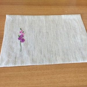 Puede incluir: Un mantel individual de lino blanco con un peque&ntilde;o dise&ntilde;o de flor bordado en tonos rosa y morado.