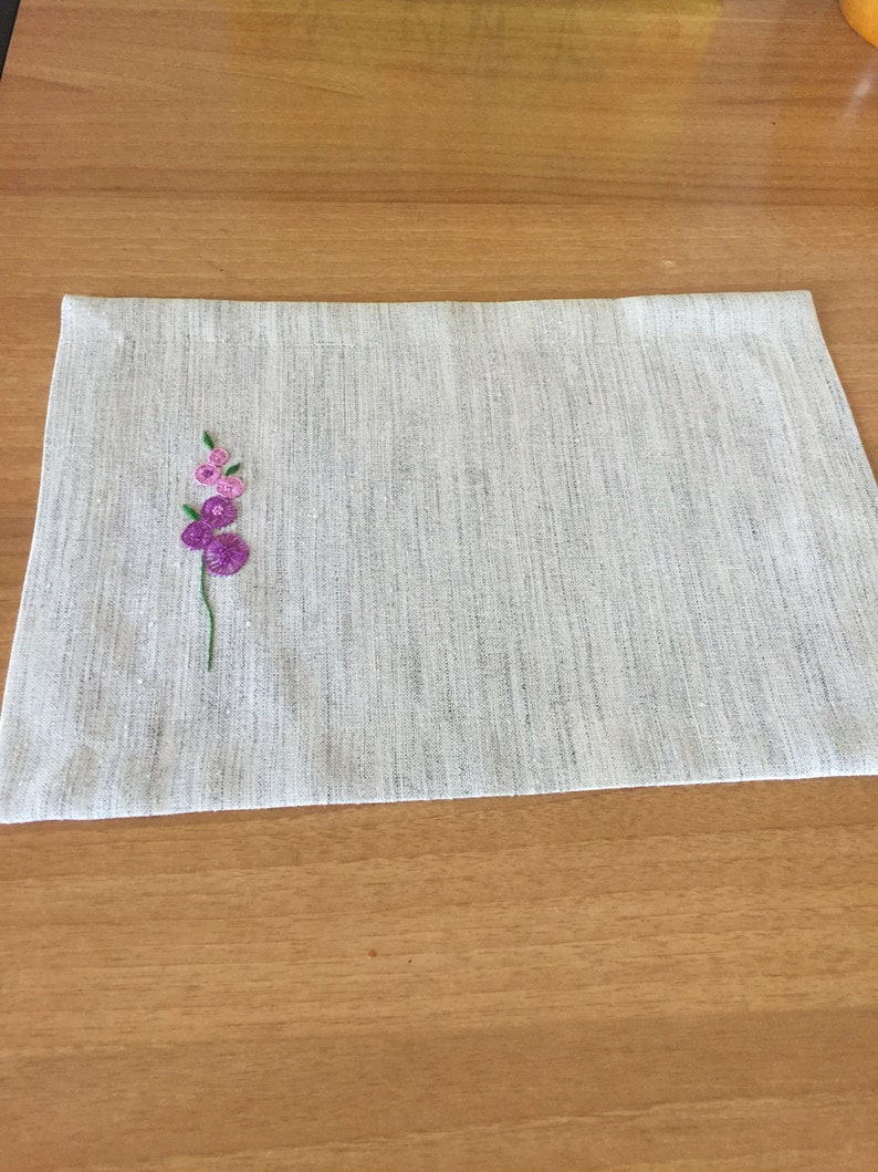 Puede incluir: Un mantel individual de lino blanco con un peque&ntilde;o dise&ntilde;o de flor bordado en tonos rosa y morado.