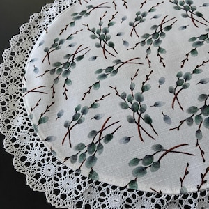May include: A round white table runner with a delicate lace trim and a green floral pattern. The pattern features small sprigs of leaves and berries.