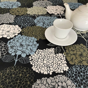 Könnte beinhalten: Eine schwarze Tischdecke mit einem weißen, blauen und grünen Blumenmuster. Eine weiße Teekanne und eine Teetasse stehen auf der Tischdecke.