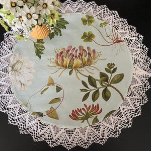 Peut inclure: Un dessous de plat rond bleu clair avec un imprimé botanique floral et une bordure en dentelle blanche. L'imprimé présente des illustrations détaillées de fleurs, de feuilles et de vignes dans des tons de vert, de rouge et de jaune. La bordure en dentelle ajoute une touche délicate.