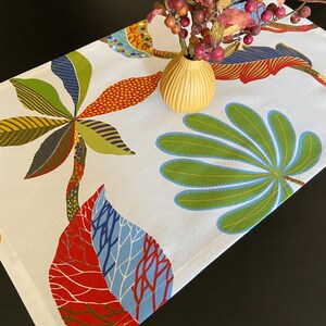 Puede incluir: Un camino de mesa blanco con un estampado floral colorido con hojas rojas, verdes, azules y amarillas. El camino de mesa está decorado con un diseño floral estilizado.