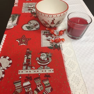 Könnte beinhalten: Ein roter Weihnachts-Tischläufer mit einem Muster aus Weihnachtsmann, Strümpfen und Geschenken, dazu eine weiße Keramikschale mit Wichtel-Illustrationen, eine rote Kerze im Glas und drei rot verpackte Bonbons auf der Stoffoberfläche.