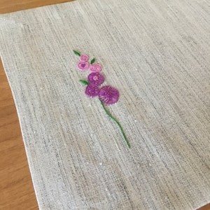 Puede incluir: Un mantel individual de lino blanco con un peque&ntilde;o dise&ntilde;o de flor bordado en tonos rosa y morado.