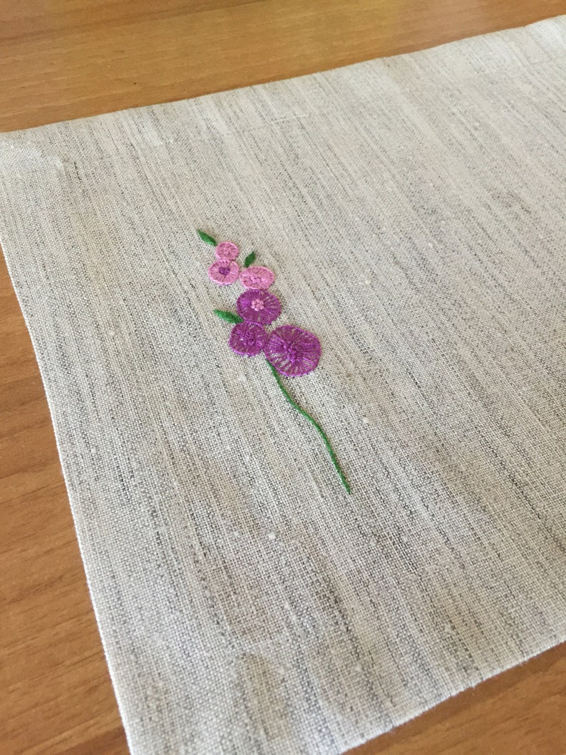 Puede incluir: Un mantel individual de lino blanco con un peque&ntilde;o dise&ntilde;o de flor bordado en tonos rosa y morado.
