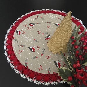 Könnte beinhalten: Ein runder Stoff-Platzdeckchen mit beigem Hintergrund, bedruckt mit rotbrüstigen Vögeln, Stechpalmenblättern und Beeren, verziert mit einer roten Spitzenborte und weißem Wellenrand. Darauf liegen ein goldener, strukturierter Baumschmuck und rote Beerenzweige.