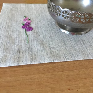 Puede incluir: Un mantel individual de lino gris con un peque&ntilde;o dise&ntilde;o de flor bordado en tonos de rosa y verde. Un cuenco de metal plateado con un dise&ntilde;o calado decorativo se encuentra sobre el mantel individual.