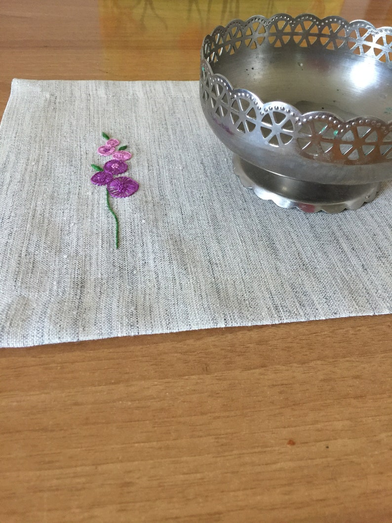 Puede incluir: Un mantel individual de lino gris con un peque&ntilde;o dise&ntilde;o de flor bordado en tonos de rosa y verde. Un cuenco de metal plateado con un dise&ntilde;o calado decorativo se encuentra sobre el mantel individual.
