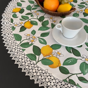 Op de afbeelding: Een ronde witte tafelkleed met een citroen- en bloemenpatroon, afgezet met een delicate kanten rand. Een fruitmand en een witte kop en schotel staan op het tafelkleed. Het tafelkleed heeft een frisse, zomerse uitstraling.