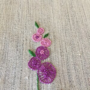 Puede incluir: Flores bordadas de color rosa y morado sobre una tela de lino beige.