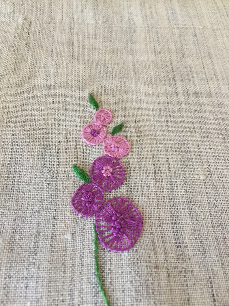 Puede incluir: Flores bordadas de color rosa y morado sobre una tela de lino beige.