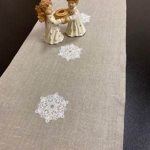 Puede incluir: Un camino de mesa beige con un patrón repetitivo de copos de nieve bordados en blanco. Dos pequeñas figuras de ángeles con túnicas color crema y detalles de estrellas doradas se sitúan sobre el camino, sosteniendo un portavelas redondo marrón.