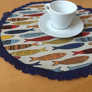 Könnte beinhalten: Eine runde Tischunterlage mit einem blauen Häkelrand zeigt ein Muster aus bunten Fischen auf weißem Hintergrund. Eine weiße Keramiktasse und -untertasse sind auf der Tischunterlage platziert.