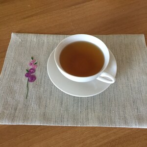 Puede incluir: Una taza de t&eacute; blanca y un platillo con t&eacute; marr&oacute;n claro en su interior, sobre un mantel individual de lino gris con un dise&ntilde;o de flor bordada de color rosa.