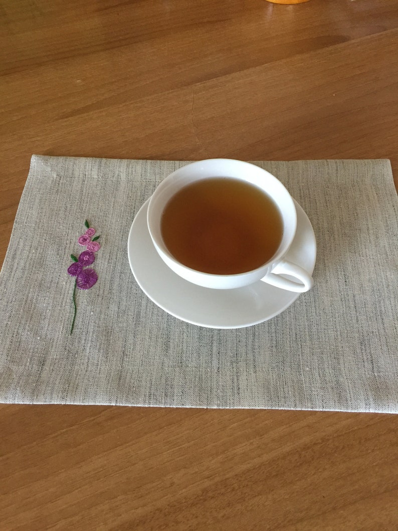 Puede incluir: Una taza de t&eacute; blanca y un platillo con t&eacute; marr&oacute;n claro en su interior, sobre un mantel individual de lino gris con un dise&ntilde;o de flor bordada de color rosa.