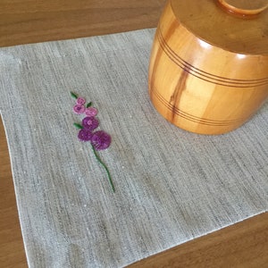 Puede incluir: Un mantel individual de lino blanco con un dise&ntilde;o de flor bordada en morado. Un recipiente de madera en forma de barril con tapa se encuentra sobre el mantel individual.