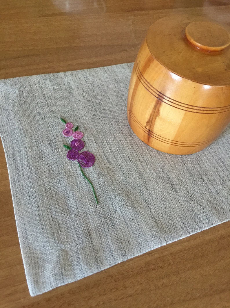 Puede incluir: Un mantel individual de lino blanco con un dise&ntilde;o de flor bordada en morado. Un recipiente de madera en forma de barril con tapa se encuentra sobre el mantel individual.