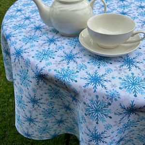 May include: White teapot and teacup with saucer on a white tablecloth with a blue snowflake pattern.