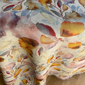 Puede incluir: Un mantel de lino con un estampado floral en acuarela. El diseño presenta flores y hojas naranjas, amarillas, rojas y moradas sobre un fondo crema. La tela está drapeada, mostrando la textura y el movimiento del material.