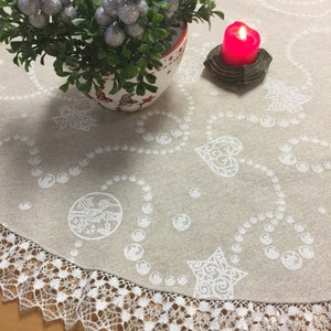 Könnte beinhalten: Ein runder, beiger Tischläufer mit einer weißen Spitzenborte. Der Läufer zeigt ein sich wiederholendes Muster aus weißen Herzen, Sternen und Ornamenten. Eine kleine Topfpflanze mit silbernen Beeren und eine rote Kerze befinden sich auf dem Läufer.