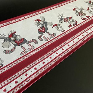 May include: A festive Christmas table runner in white and red, decorated with cartoon reindeer in hats and sweaters. The runner has a red border with snowflakes and is approximately 152 cm long.