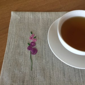 Puede incluir: Un mantel individual de lino gris con un peque&ntilde;o dise&ntilde;o de flor bordado en tonos de morado y verde. Un platillo blanco con una taza de t&eacute; se encuentra sobre el mantel individual.