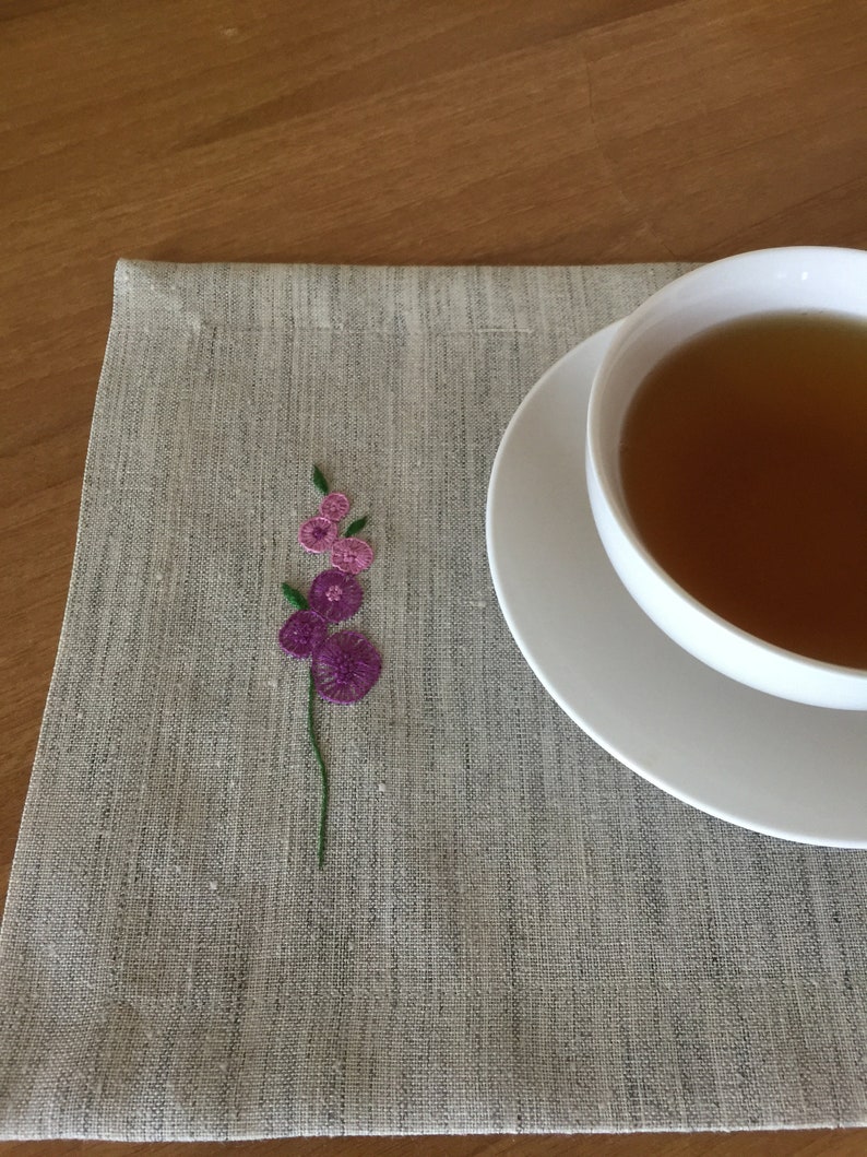 Puede incluir: Un mantel individual de lino gris con un peque&ntilde;o dise&ntilde;o de flor bordado en tonos de morado y verde. Un platillo blanco con una taza de t&eacute; se encuentra sobre el mantel individual.