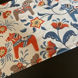 Pode incluir: Um caminho de mesa de tecido branco com um padrão repetitivo de cavalos Dala estilizados em laranja, azul e castanho, intercalados com flores cor de laranja e amarelas e videiras de folhas azuis. O tecido tem uma textura visível.