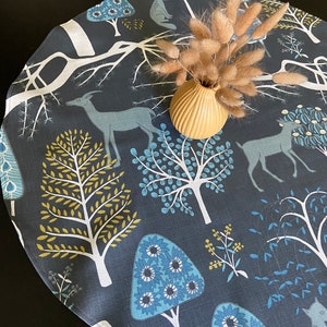 Może przedstawiać: Okrągły bieżnik na stół z ciemnoniebieskim tłem i fantazyjnym wzorem lasu. Wzór przedstawia stylizowane drzewa, jelenie i lisy w odcieniach niebieskiego, zielonego i żółtego.